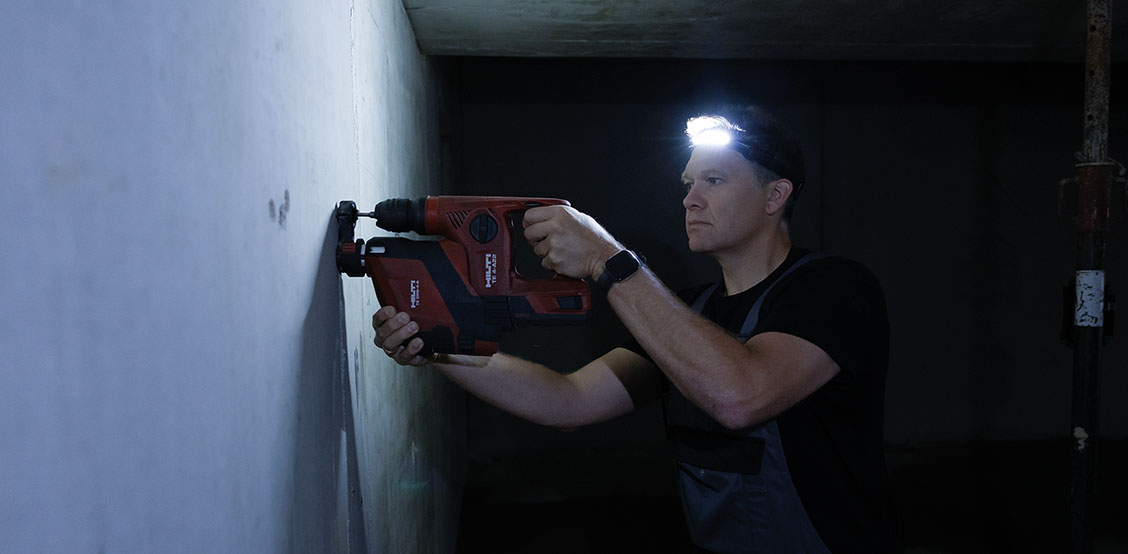 Un técnico con una linterna de cabeza STAHLWILLE taladra un agujero en una pared con un martillo rotativo en condiciones de poca luz
