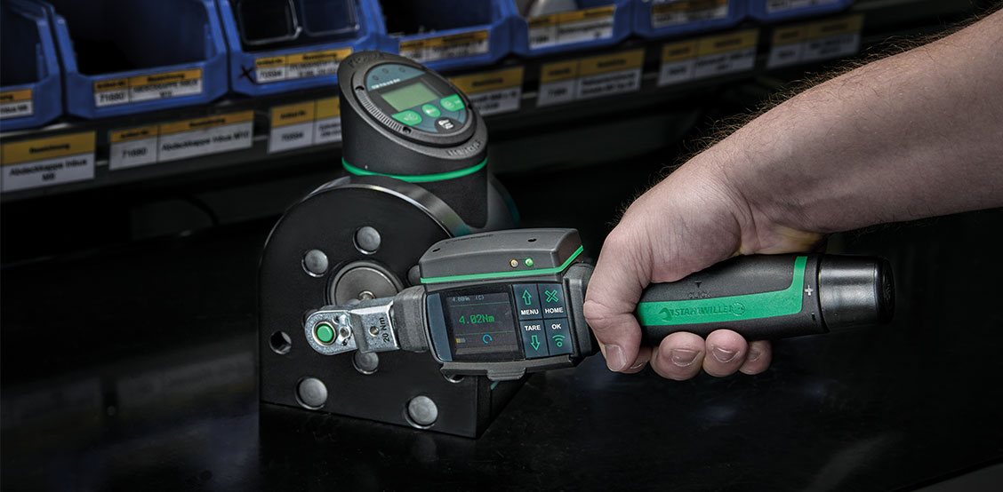 Close-up of a STAHLWILLE torque wrench MANOSKOP 766 DAPTIQ with WLAN module during assembly of a tester. Measured values can be seen on the screen, while workshop shelves and organiser boxes in the background make clear that the application relates to precise torque measurement and quality assurance.